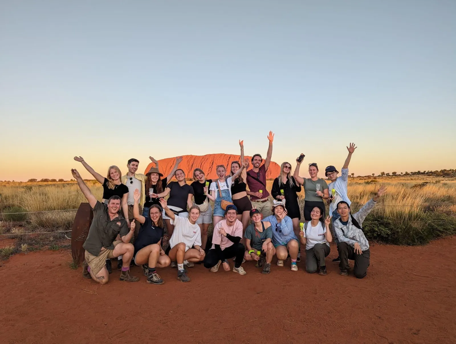 Large tour group cheering with glasses of sparkling wine on red ground, Uluru lit orange behind them