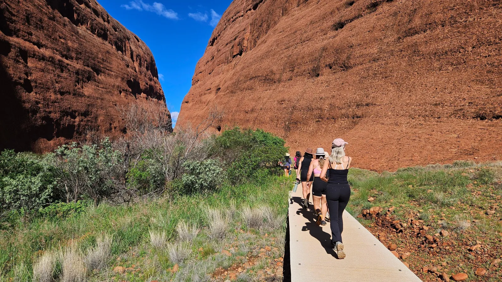 Hikers in single file on a raised boardwalk leading into a narrow gorge between Kata Tjuta's red walls
