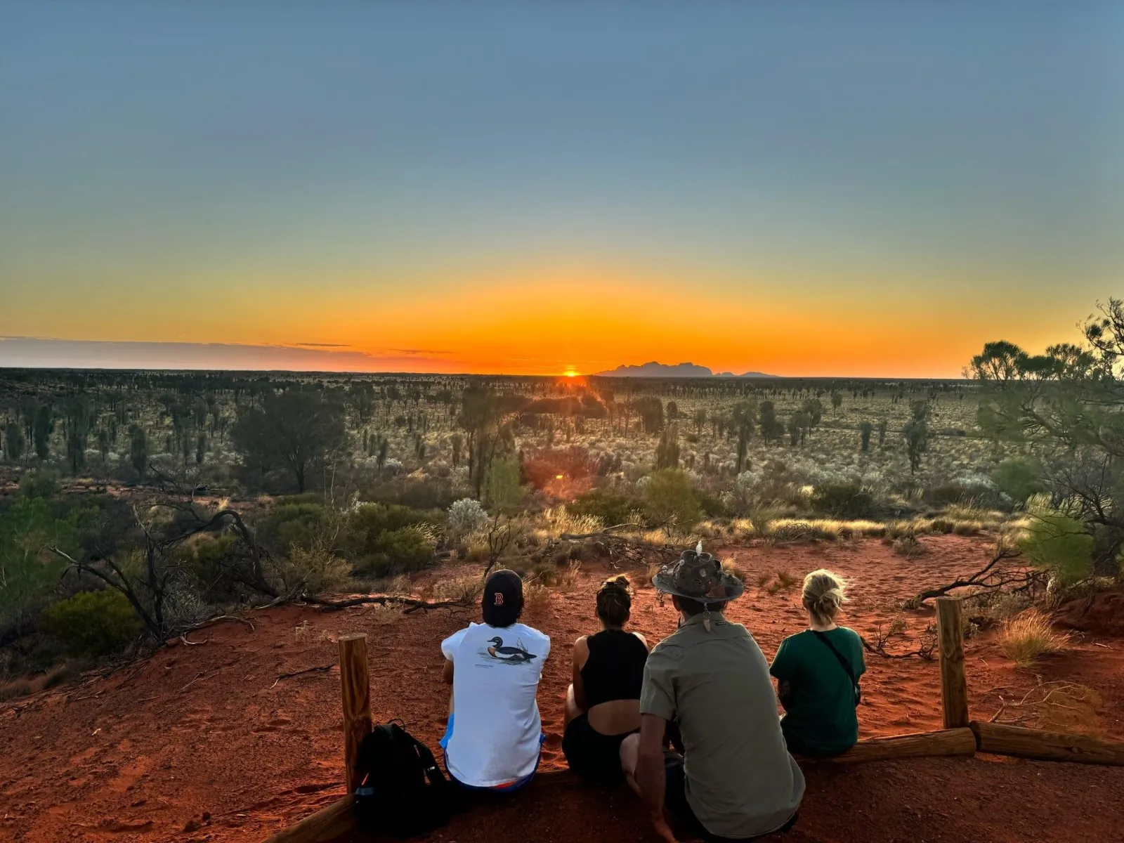 Four people on a dune lookout watching the sun drop beside the silhouette of Kata Tjuta