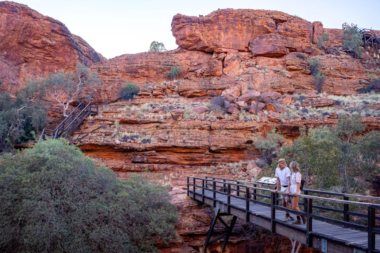 Couple on a wooden footbridge below stepped red sandstone cliffs and a staircase at Kings Canyon