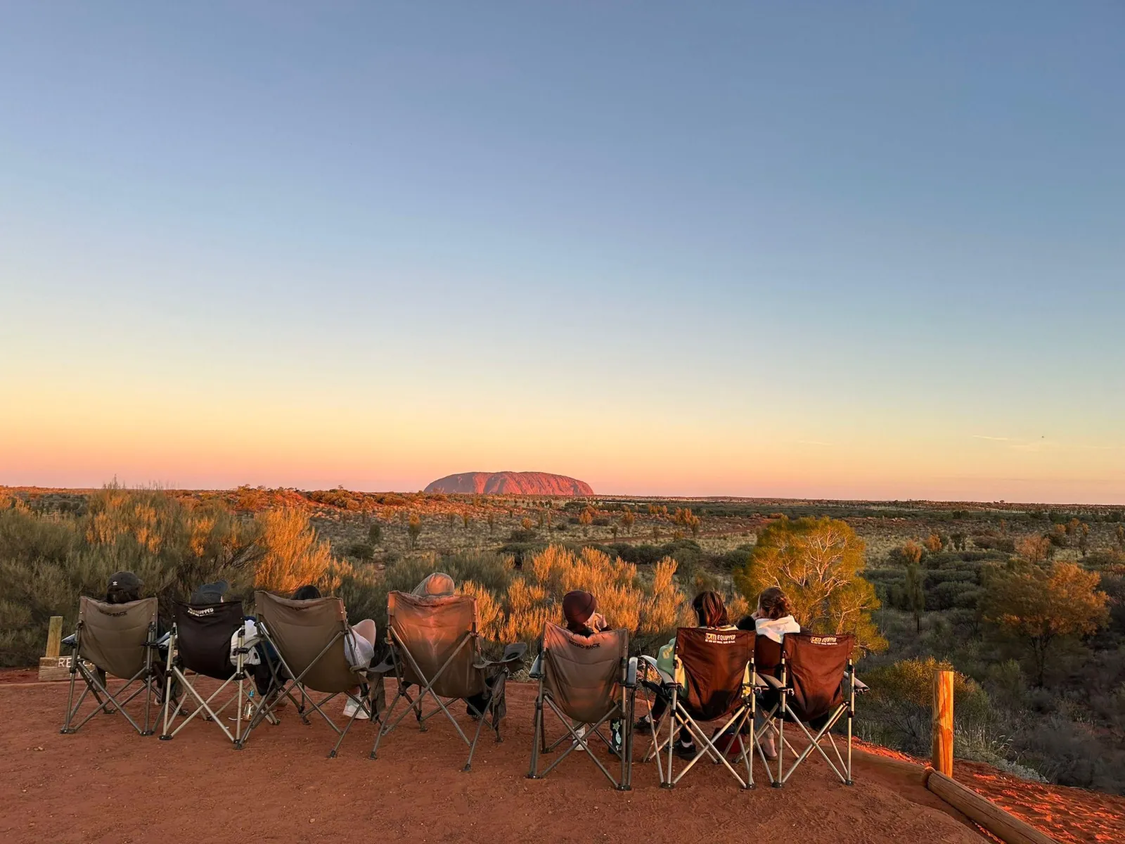 Row of travelers in camp chairs on a red dune watching Uluru in golden low-angle light