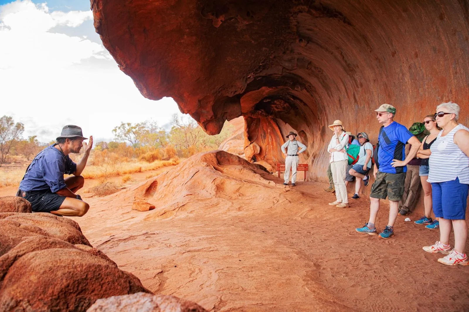 Guide crouching to talk to a tour group standing under a wave-shaped rock overhang at the base of Uluru