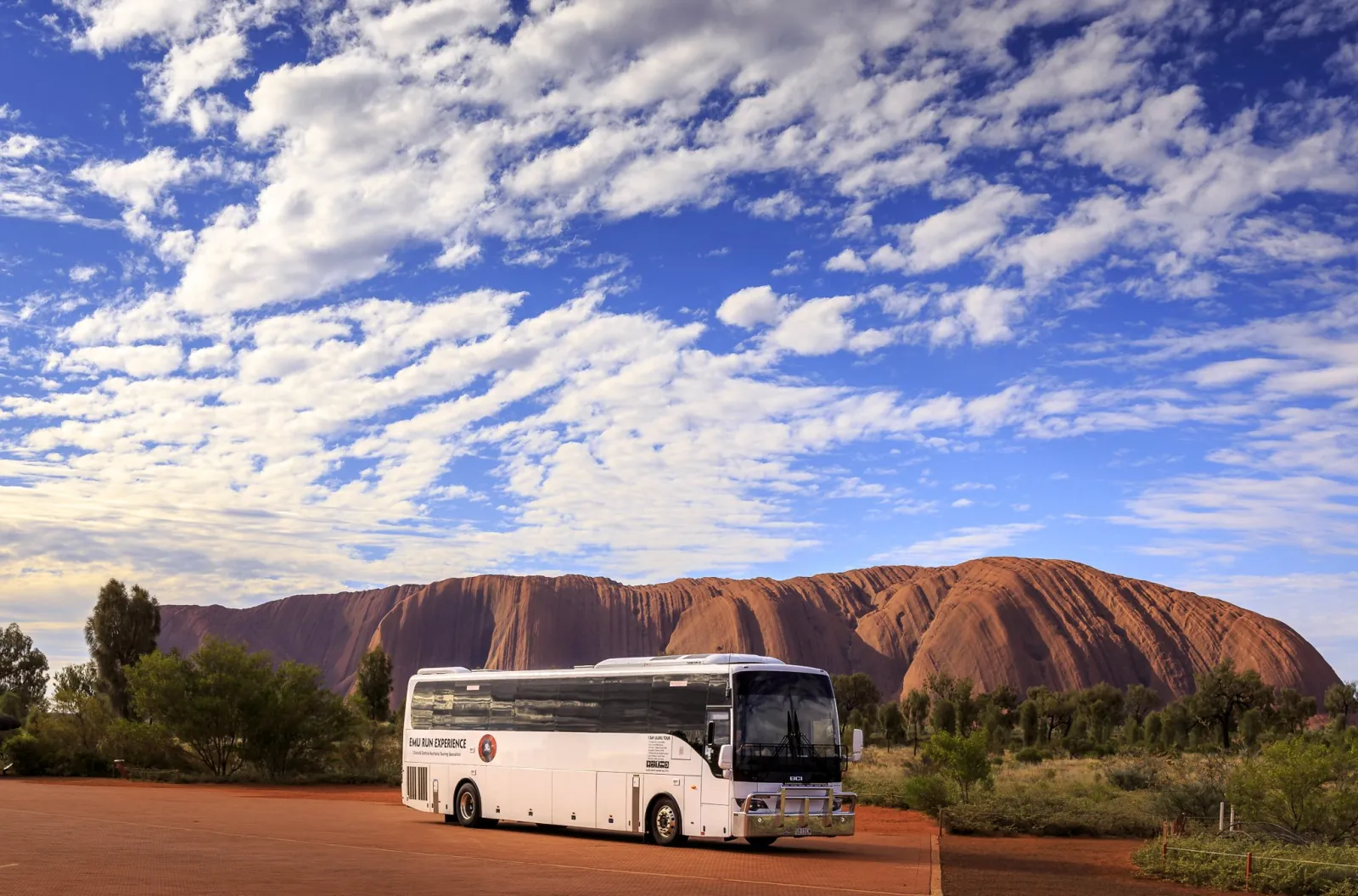 White Emu Run Experience coach parked on red ground with Uluru and a cloud-streaked blue sky behind