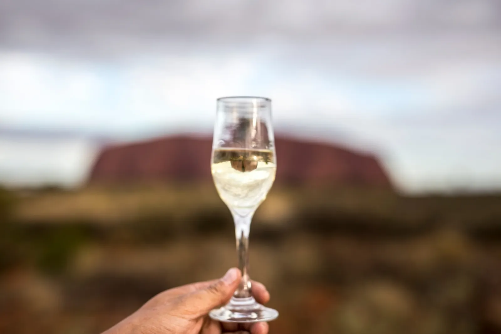 Hand holding a glass of sparkling wine in front of a softly blurred Uluru at dusk
