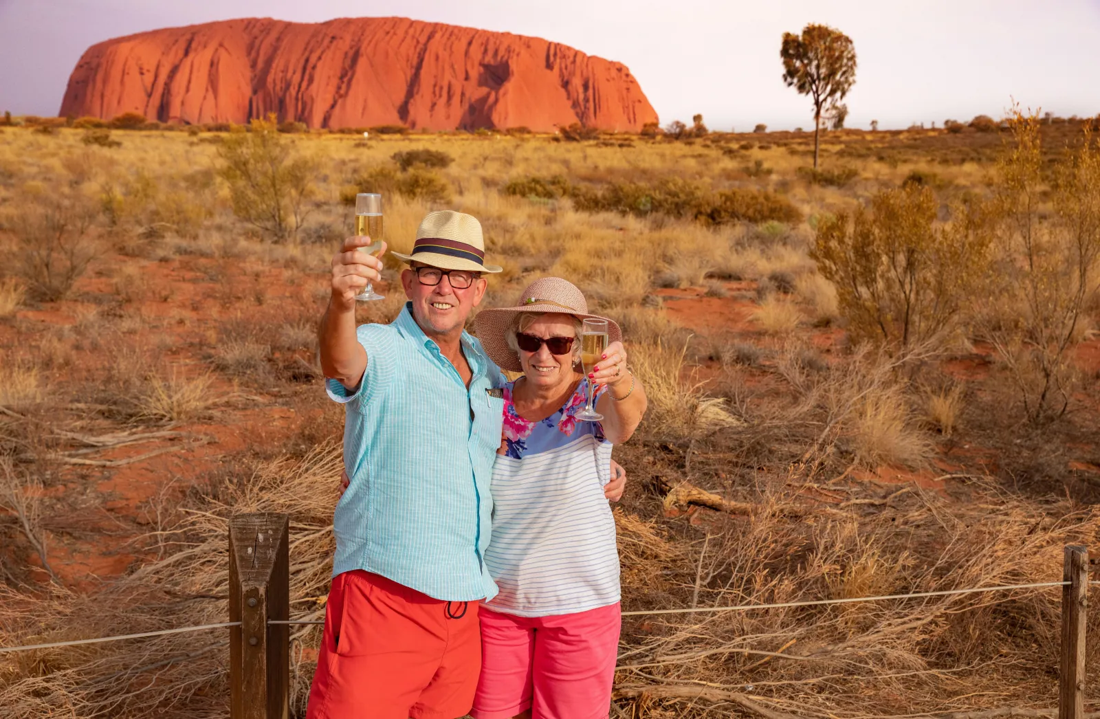 Couple raising glasses of sparkling wine at a fenced viewing area with Uluru glowing red behind them