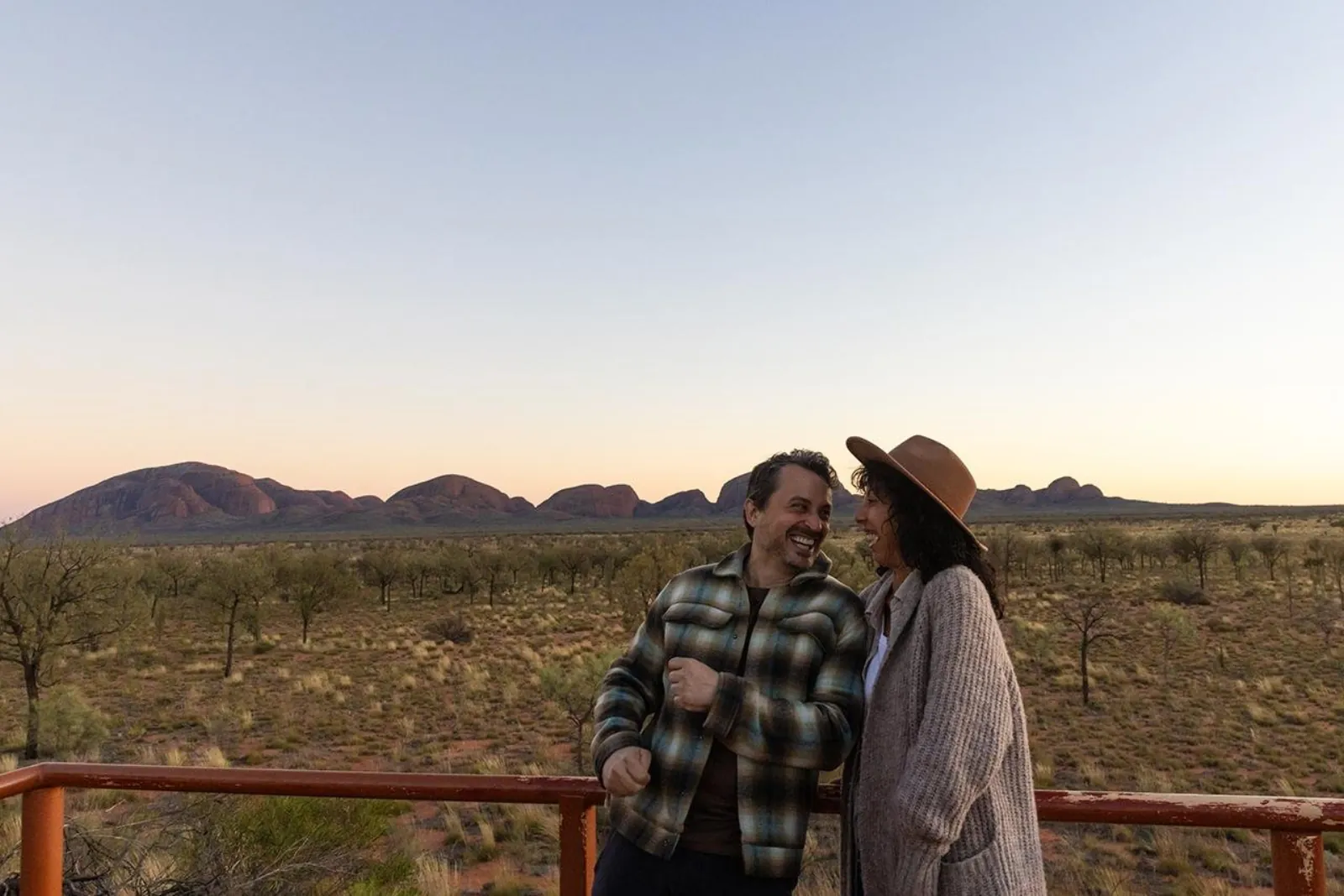 Couple laughing at a viewing platform railing with the long row of Kata Tjuta domes behind