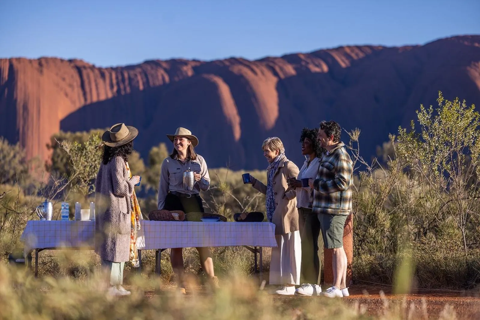 Guide serving hot drinks from a checked-cloth table to guests near the lit face of Uluru