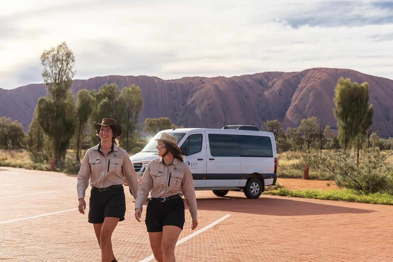 Two SEIT guides walking across a red car park with their white small-group van and Uluru behind