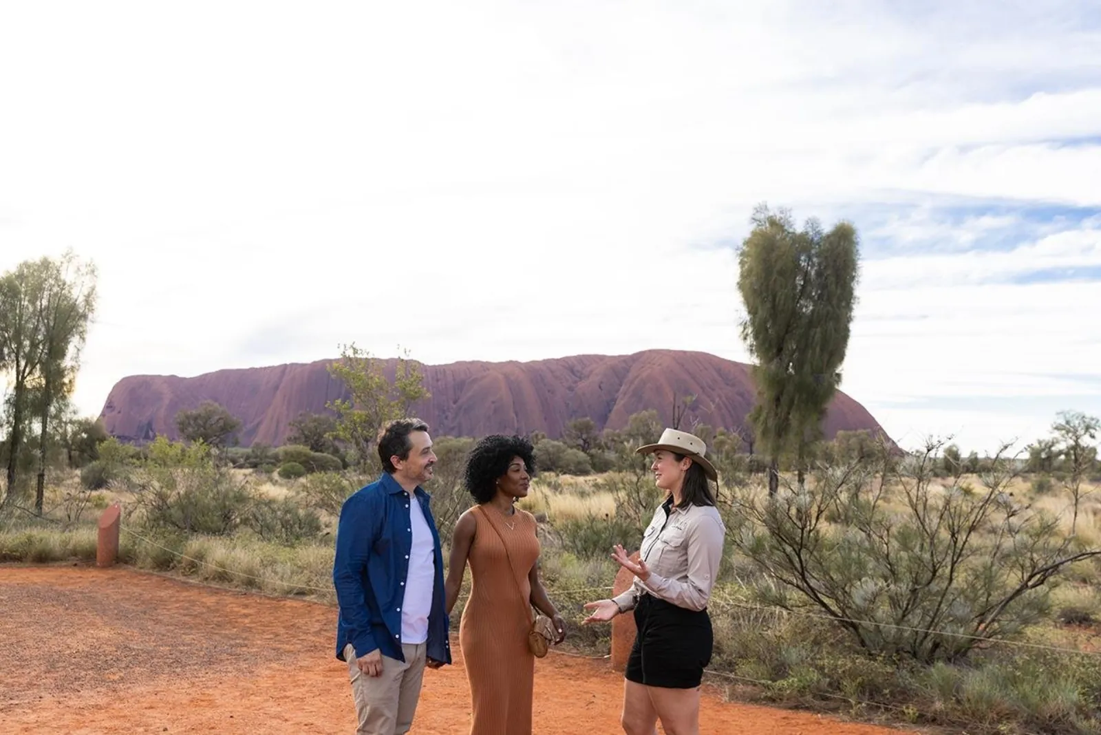 SEIT guide in a bush hat talking with a couple on a red dirt path with Uluru in the distance