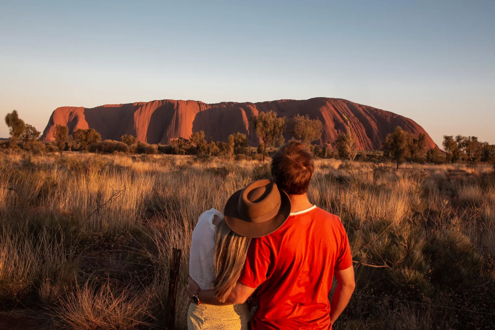 Couple standing in tall grass watching late light on Uluru's long red face