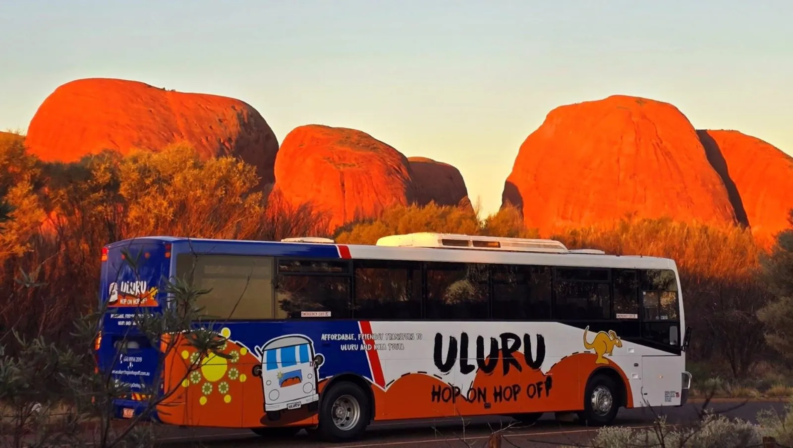 Uluru Hop On Hop Off bus on the road below the red domes of Kata Tjuta lit by low sun
