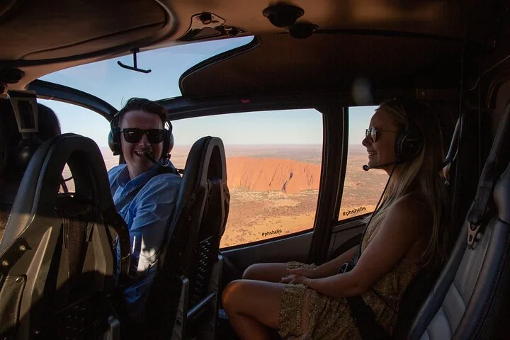 Passengers in headsets inside the helicopter cabin with Uluru beyond the window