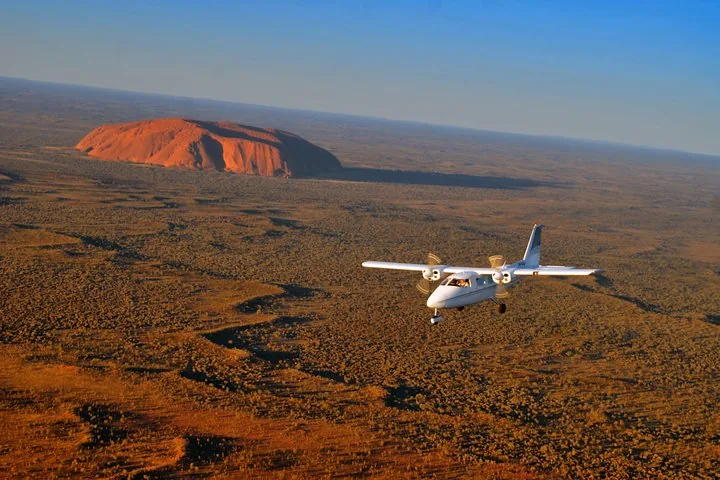 White twin-engine plane flying over red desert with Uluru on the horizon in low light