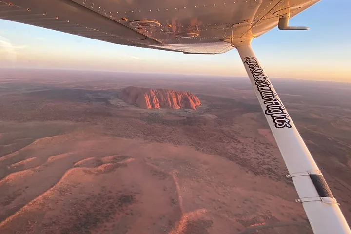 View under a high wing over sand ridges toward Uluru in soft evening light