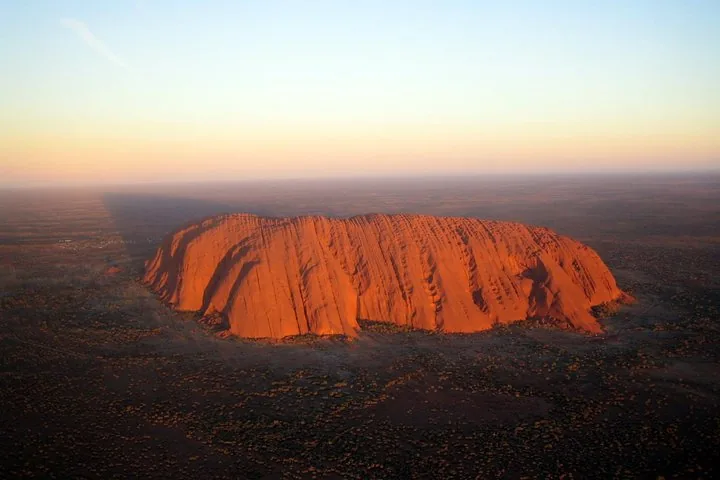 Uluru seen from the air at dusk, its long face lit orange against a darkening plain
