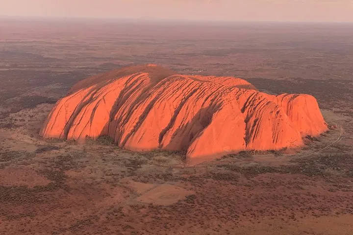 Uluru from the air in soft pink light, its ribbed red sides rising from the flat plain