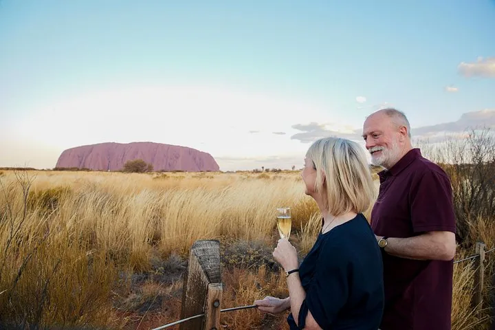 Couple with sparkling wine at the viewing area fence as Uluru turns pink across golden grass