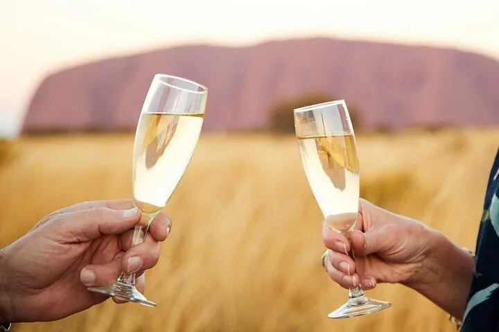Two glasses of sparkling wine raised in a toast with a blurred pink Uluru behind
