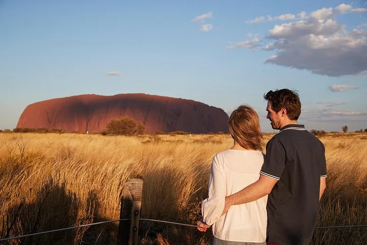 Couple at a wooden rail looking across windblown grass at Uluru in late-afternoon light