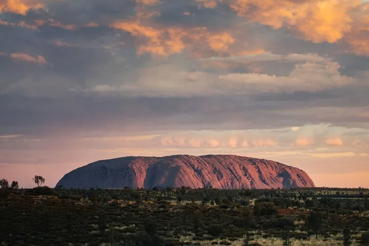 Uluru lit soft pink beneath layered orange and grey sunset clouds over dark scrub
