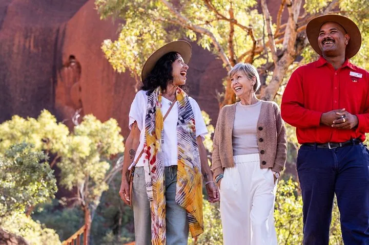 Guide in a red shirt and hat laughing with two guests beside trees at the foot of Uluru