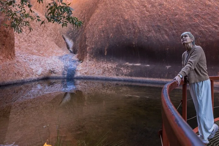 Visitor at a railing looking up at the dark water streak above a waterhole at the base of Uluru