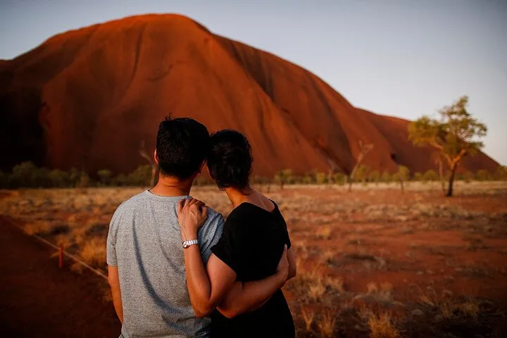 Couple standing arm in arm looking at Uluru lit deep red in the early morning light