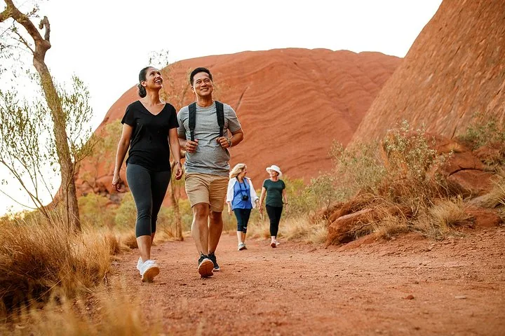 Group of four walkers on a red dirt path beside the curved sandstone walls of Uluru
