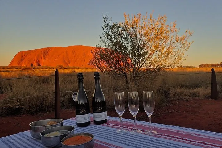 Sparkling wine bottles, glasses and dips on a table as Uluru glows orange at sunset behind