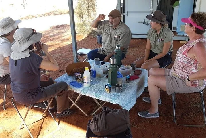 Hosts and guests sharing tea from thermoses at a folding table set up on red sand