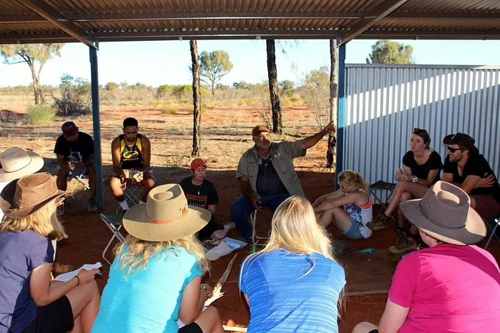 Aboriginal host speaking to a seated group under an open-sided shelter on red ground