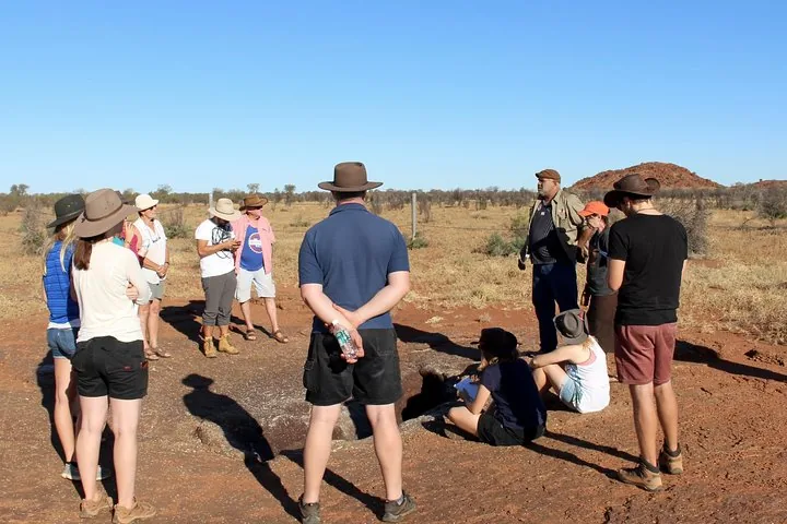 Group in sun hats standing around a rock hole in open scrubland, listening to their Aboriginal host