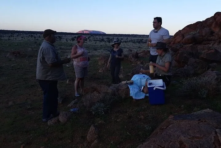 Guests with drinks among boulders at dusk, a cooler at their feet and Uluru pink on the horizon